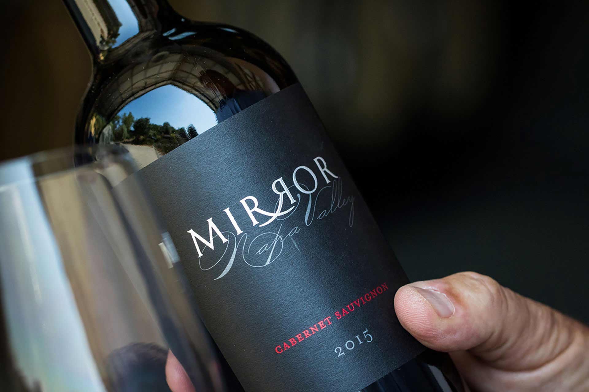 Close-up of a hand holding a Mirror Cabernet Sauvignon 2015 wine bottle with a reflective glass.