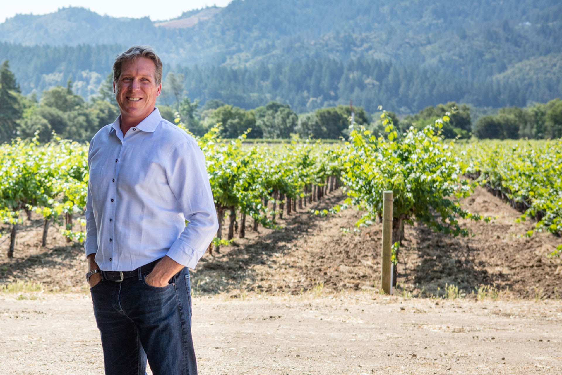 Man standing in vineyard with lush green vines and mountains in the background.
